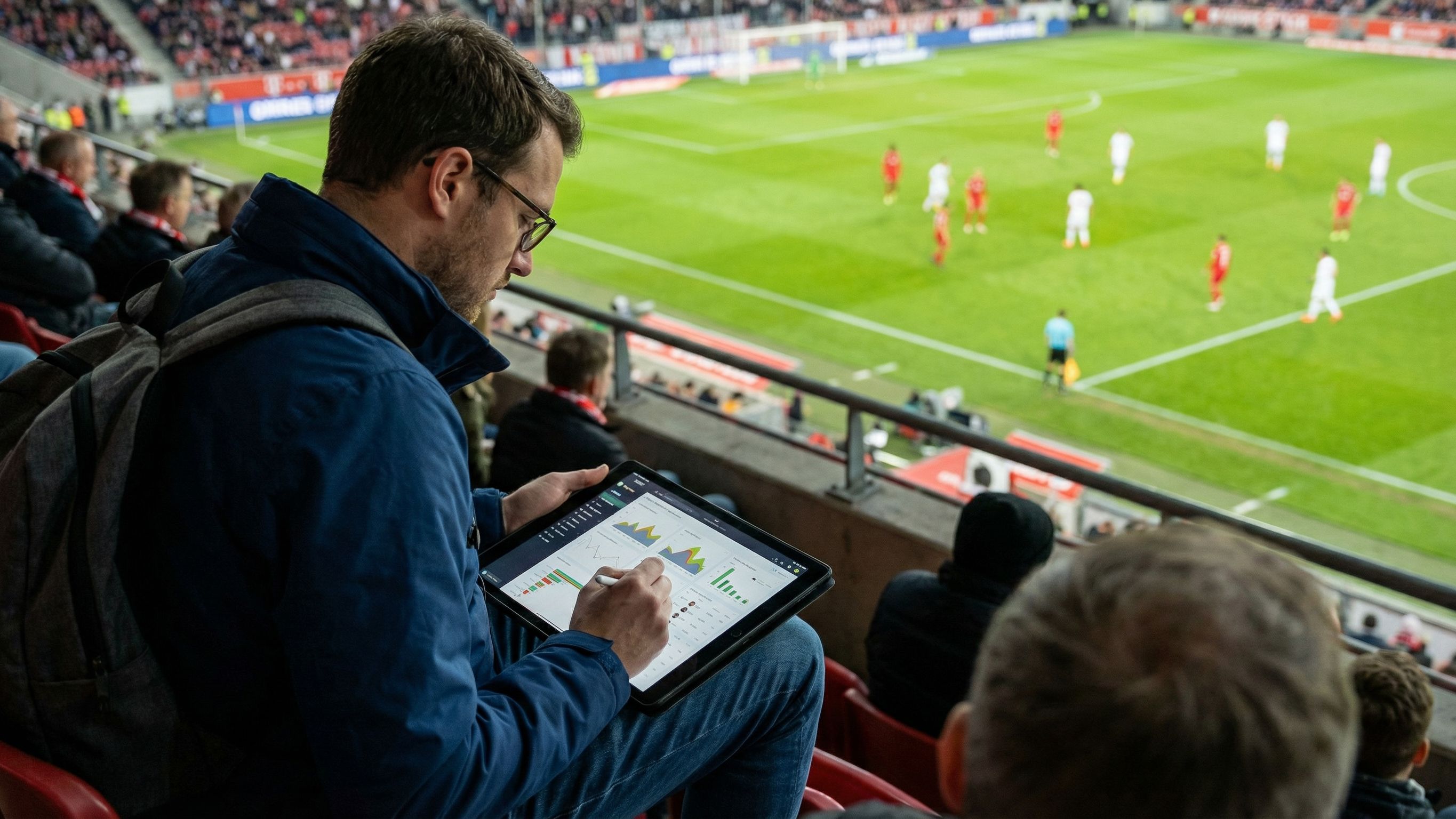A sports performance analyst sits on the sidelines of a football match, holding a tablet displaying a colourful heatmap while reviewing a laptop screen showing generic data dashboards with bar graphs, line charts, and analytics widgets. Players and the pitch are blurred in the background.