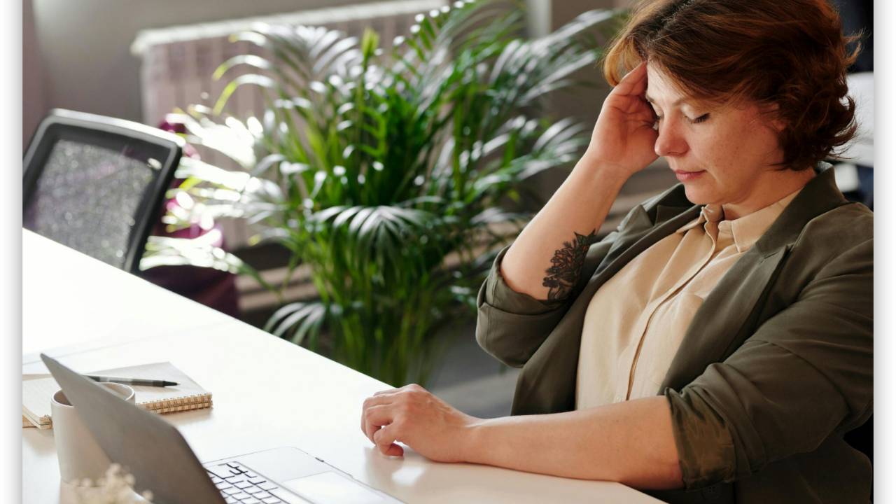 Fair skinned woman sits at a desk with a laptop, tucking her short brown hair behind her ear, looking pensive