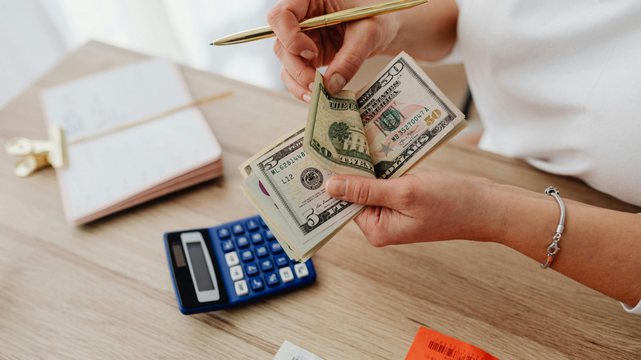 A close up of a pair of hands counting a stack of bills over a desk with a notebook and a calculator.