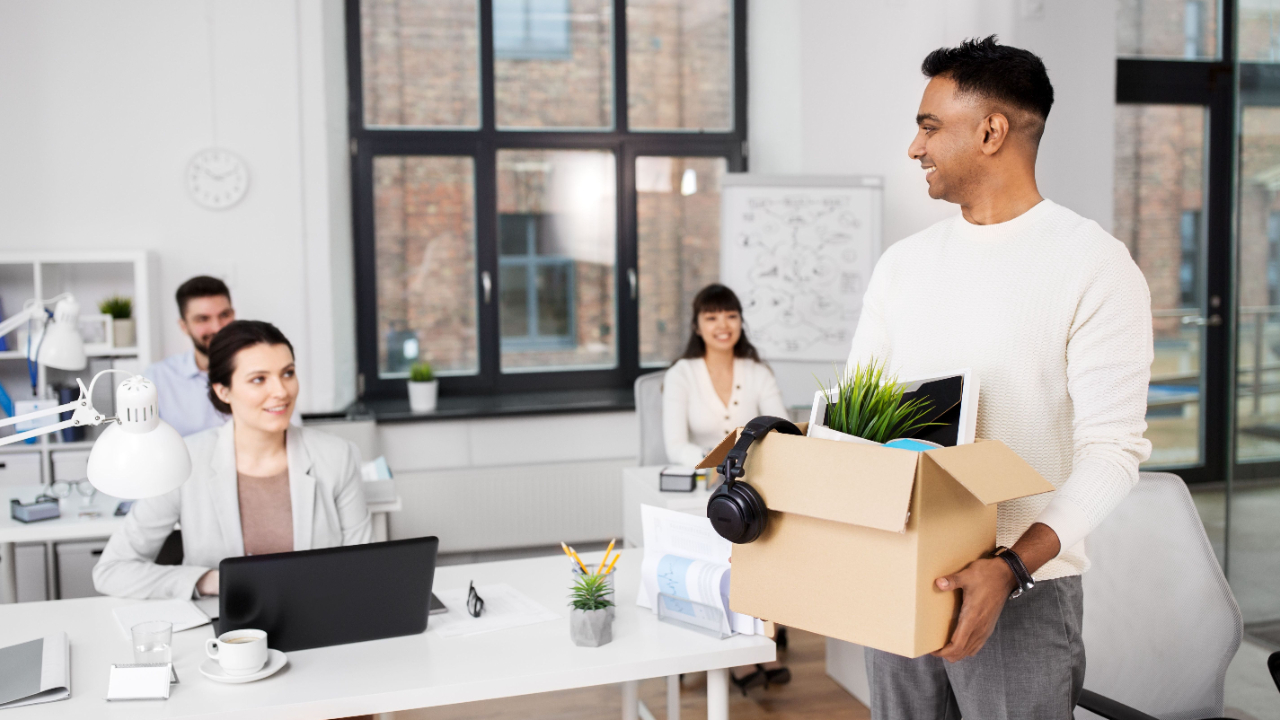 Employee leaving office carrying box of personal belongings while coworkers look on