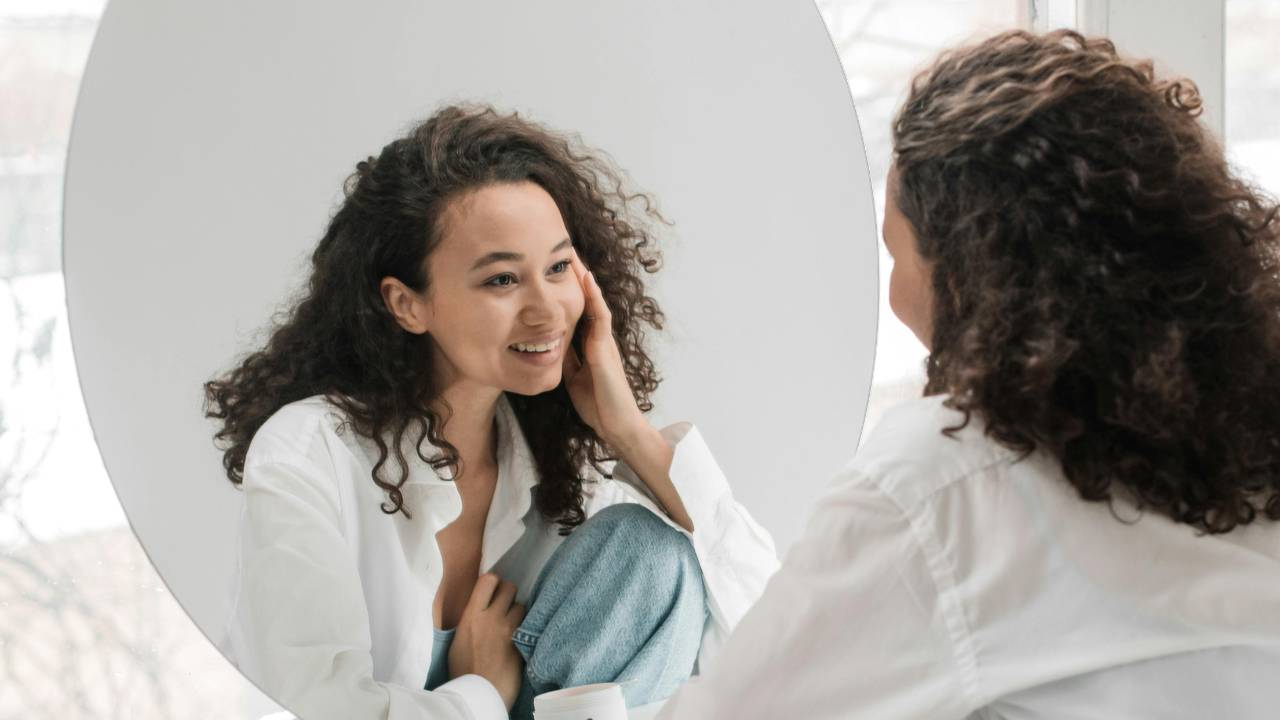 Woman smiling softly in mirror, symbolizing self-compassion, healing, and freedom from self-criticism.