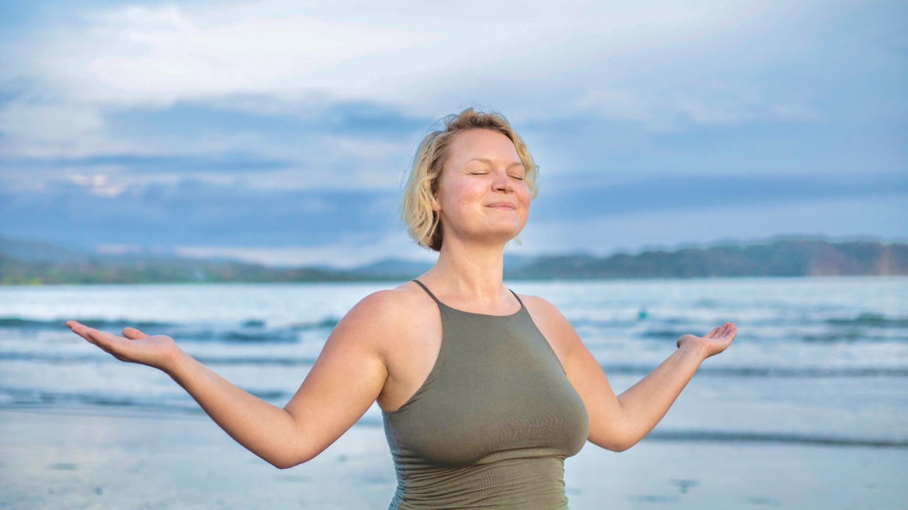 Woman near ocean smiling with eyes closed and arms slightly lifted at her sides, symbolizing peace, clarity, and the freedom that comes from letting go of control.