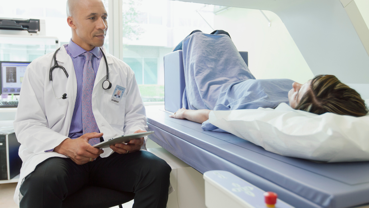 a woman lying down on a DXA scan with a doctor sitting next to her holding a clip board