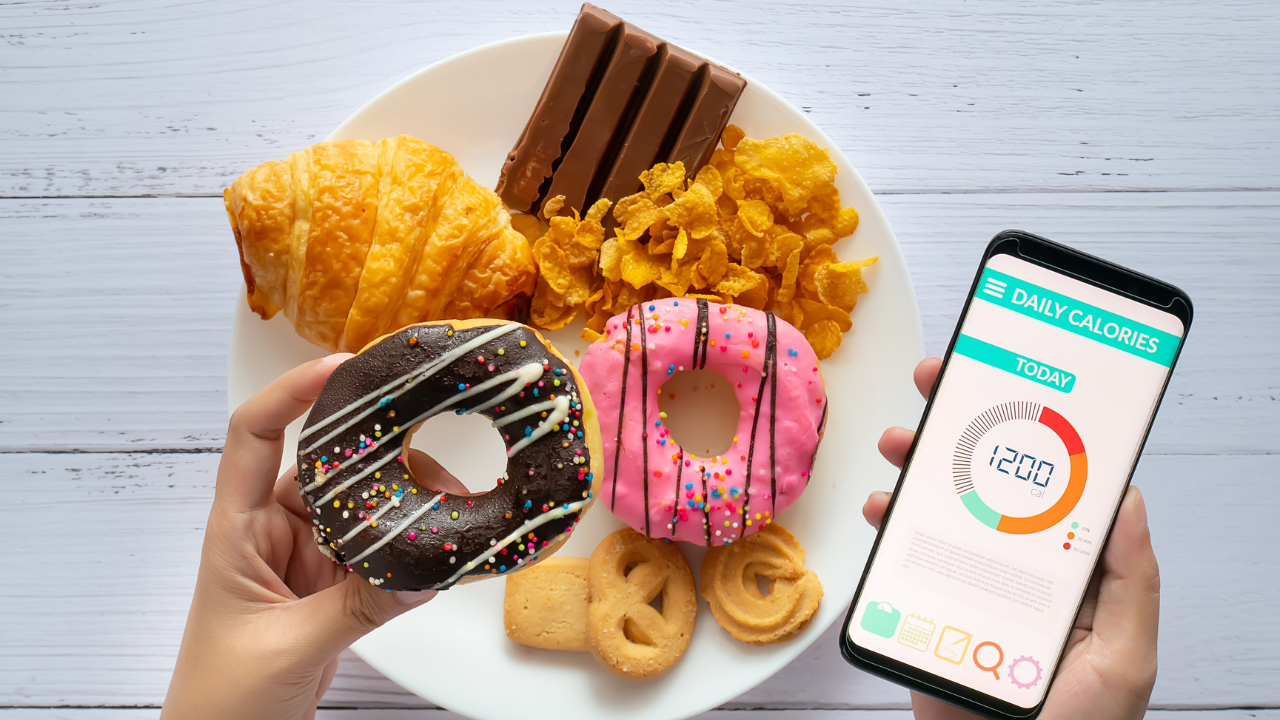 a hand holding a donut next to another hand holding a phone with a macronutrient tracking app opened, all in front of a plate of donuts and other junk food
