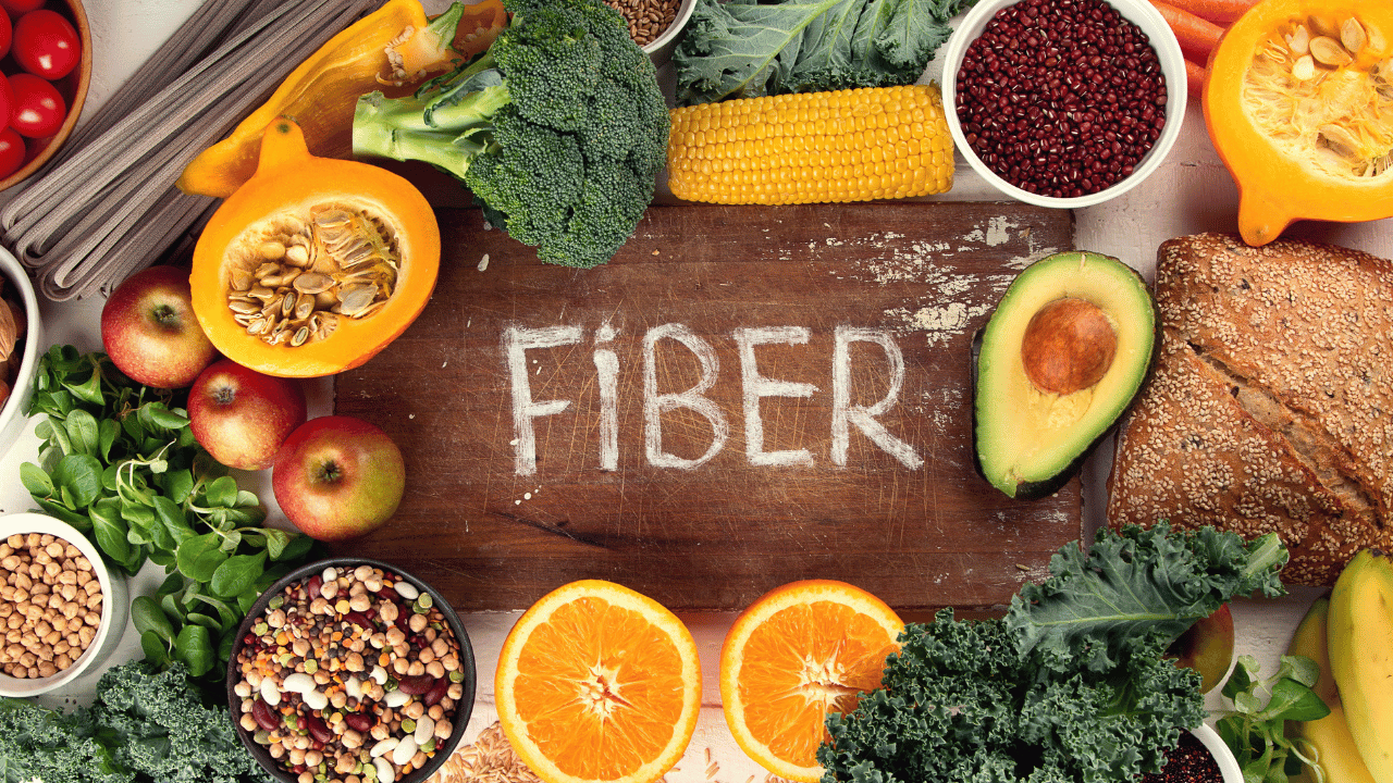 The word fiber written on a cutting board in white chalk surrounded by a variety of colorful high fiber fruits, veggies, and whole grains