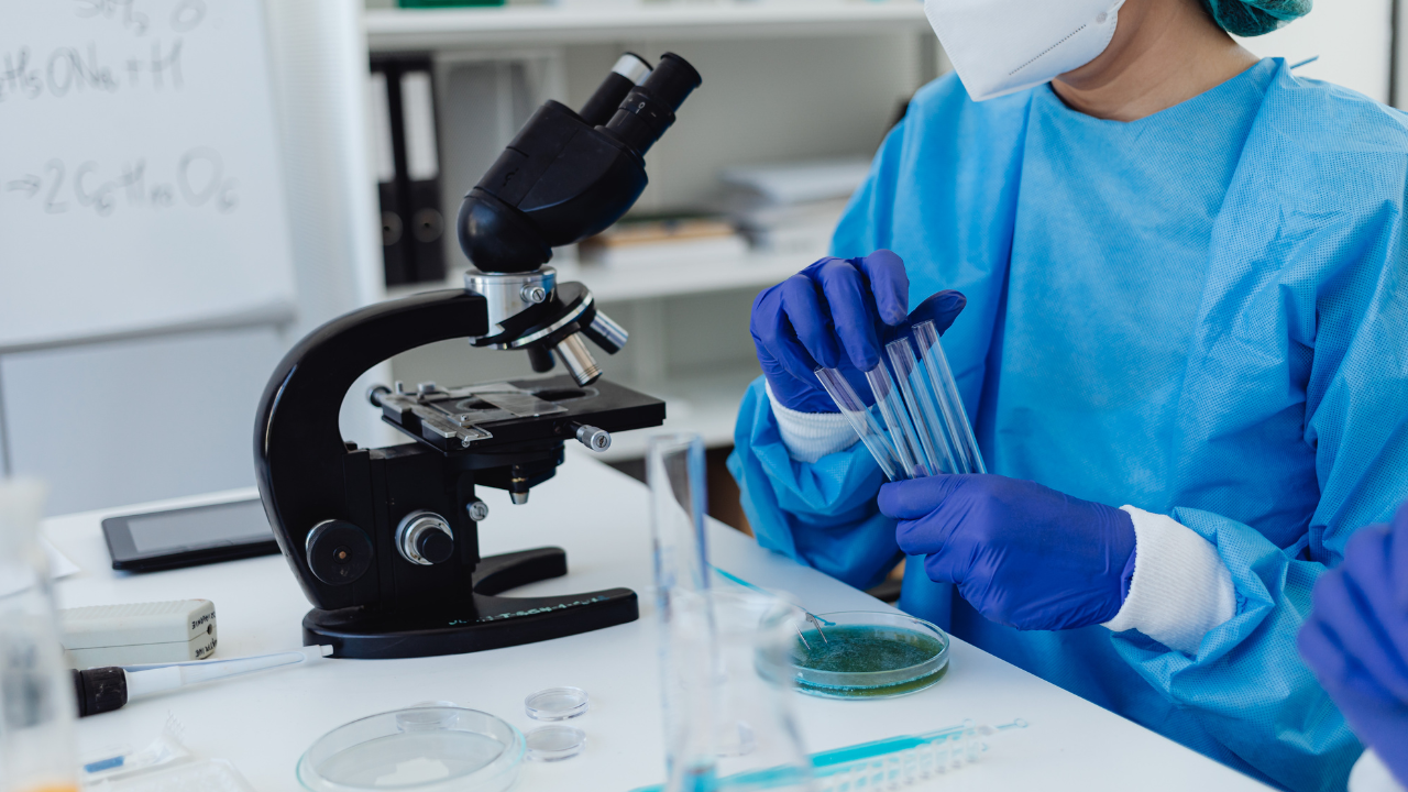 women in a blue chem suit with test tubes in her hand and a microscope on the table