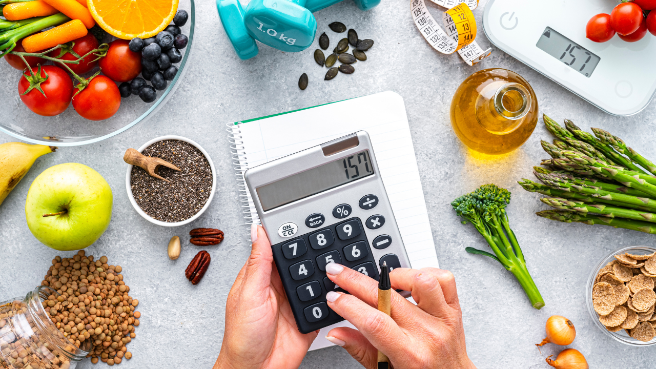 two hands holding a calculator in front of a notepad surrounded by a large variety of healthy foods, a tape measure, and a food scale