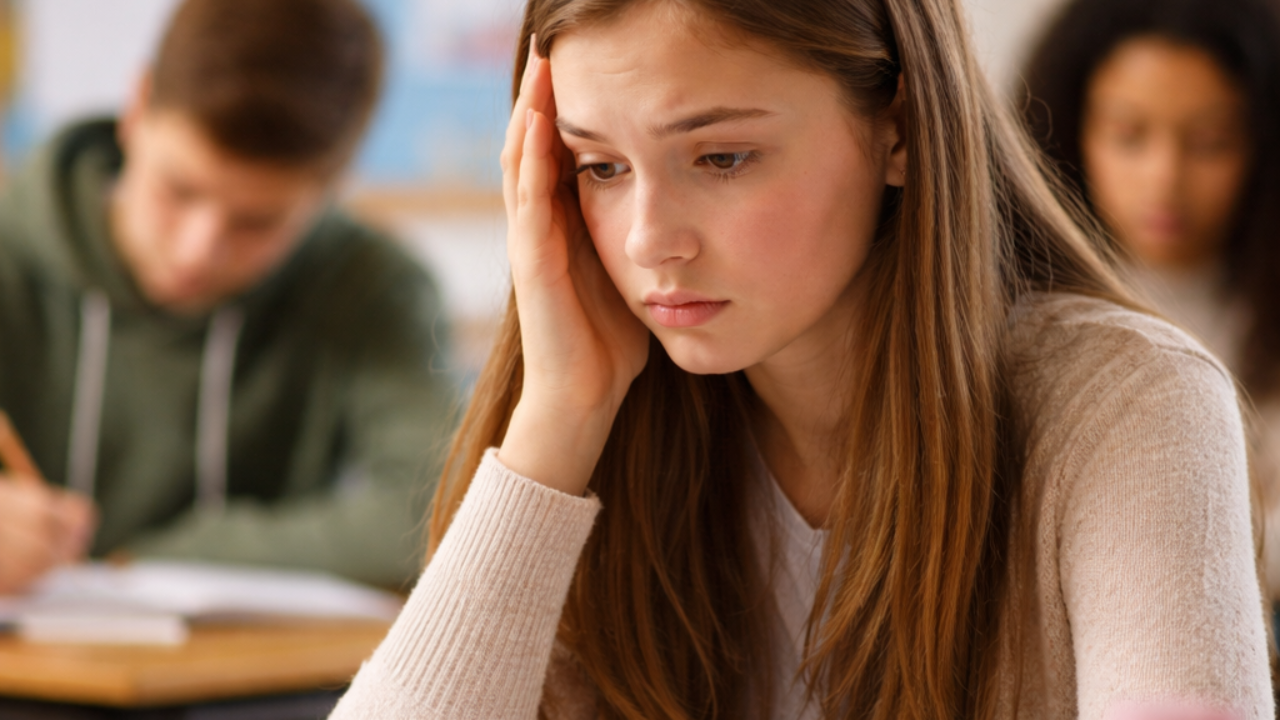 Teenage girl sitting in a classroom looking anxious during an exam, holding her head and appearing stressed.