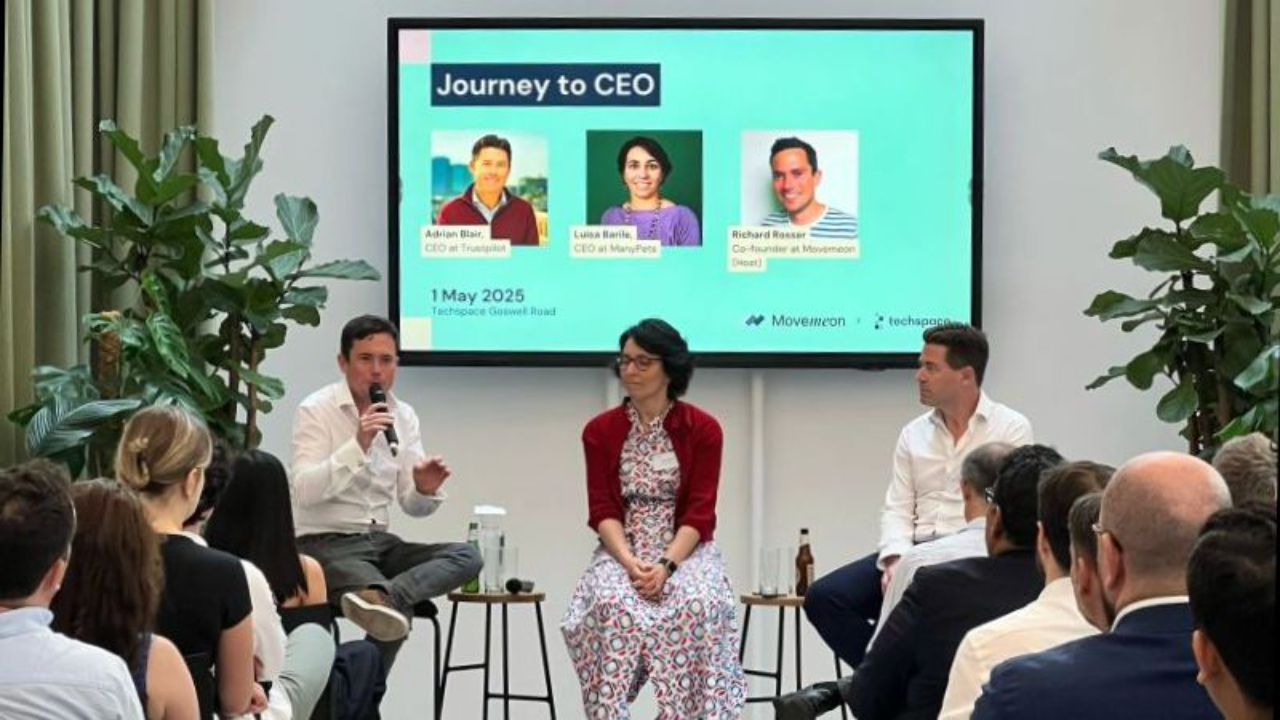A three-person panel sits on stools in front of a large screen, speaking to a full seated audience on the topic of Path to CEOin a bright room with tall curtains and plants.