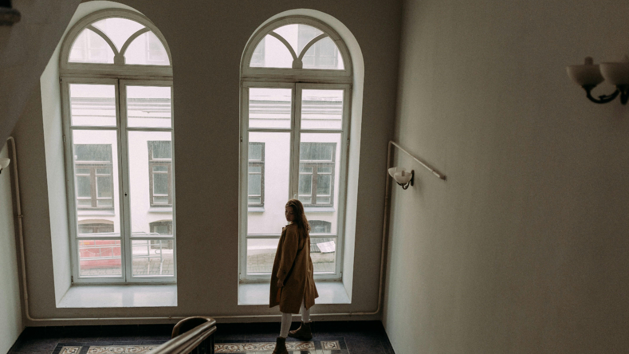 Professional woman standing by tall windows in a quiet, light-filled building, reflecting.