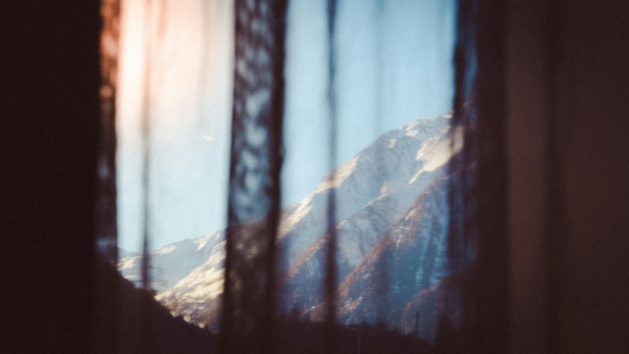 Snow-covered mountain seen through a softly blurred window, with warm indoor light reflecting on the glass.