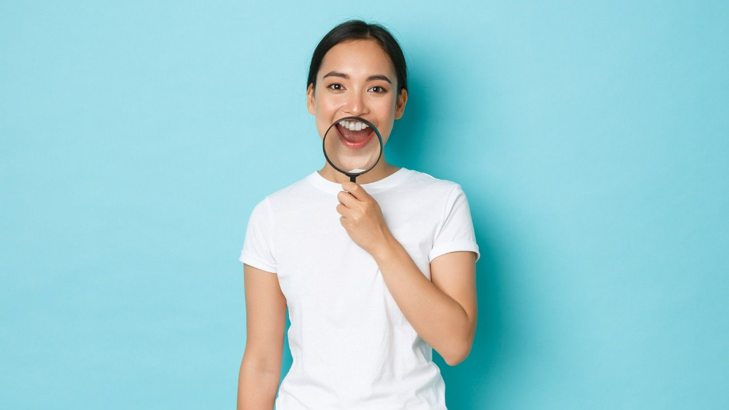 Woman examining oral health and chronic disease connection with magnifying glass on teal background.