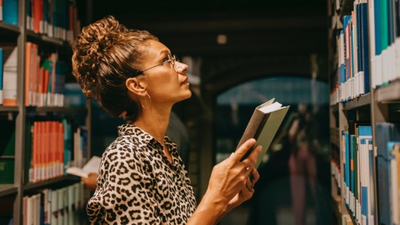Coach selecting a book from a library shelf while researching professional development and coaching psychology.
