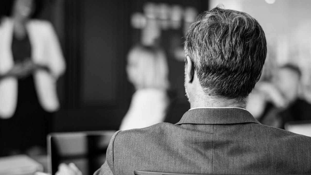 A black and white, rear-view shot of a man in a grey suit sitting in a boardroom. He is looking towards a blurred presenter at the front of the room, representing the "Connection Debt" and the gap between leadership and team alignment.