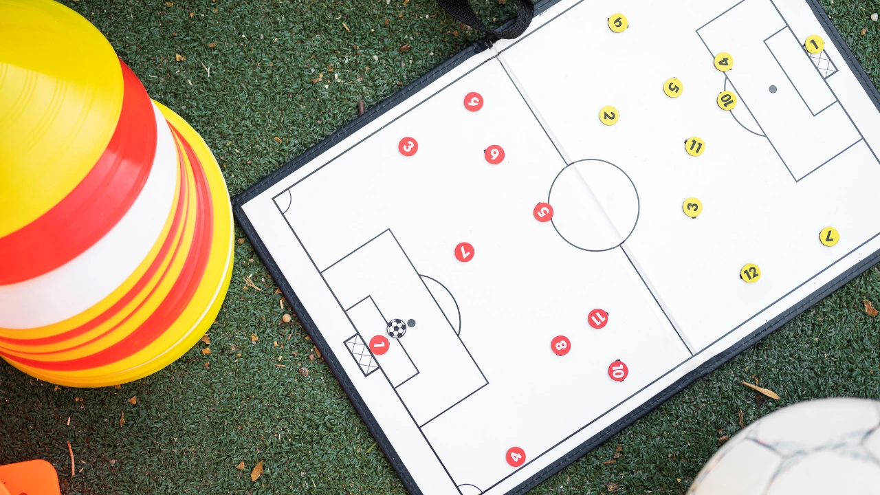 A high-angle shot of a coach's tactical whiteboard showing a football (soccer) pitch layout with red and yellow player markers, resting on green astroturf next to a stack of yellow training cones and a white football to represent a leadership teams formation 