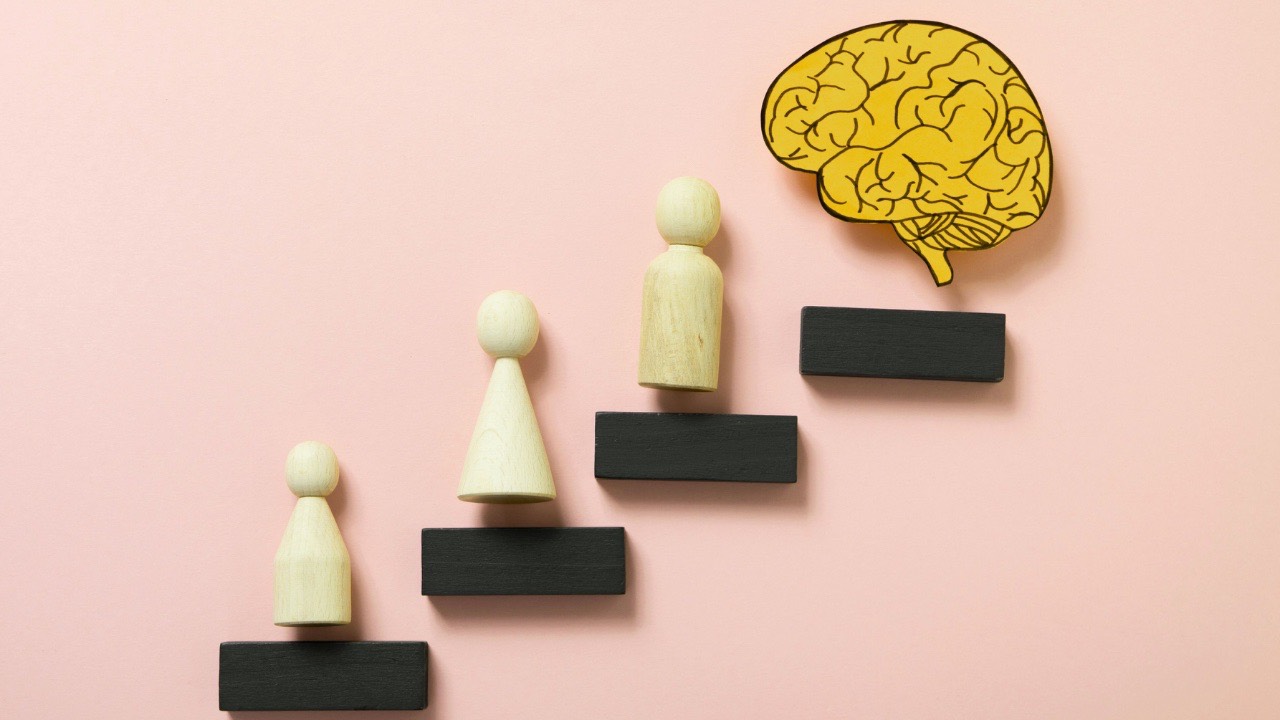 Wooden human figures climbing black steps toward a yellow brain symbol, representing leadership growth, emotional intelligence, and understanding the limits of the human brain.
