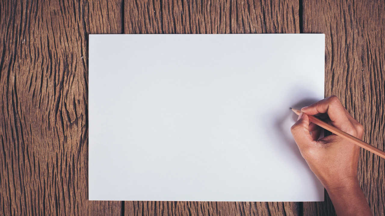 Hand holding a pencil over a blank sheet of paper on a wooden table, ready to write.
