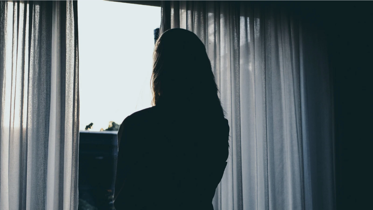 A silhouetted figure standing near a window with sheer curtains, light filtering into a quiet interior.