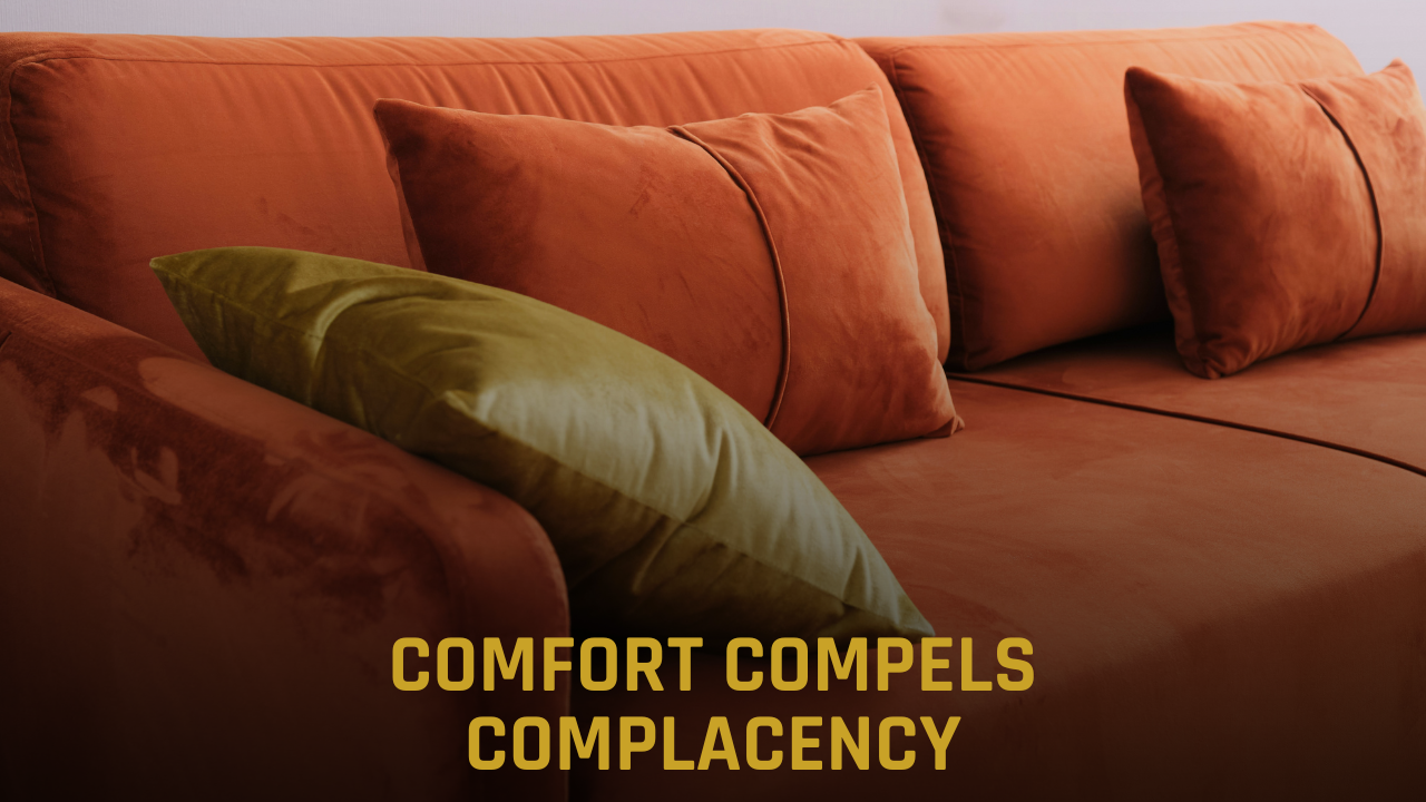 A moody, high-contrast photo of a plush orange-red couch with several soft pillows, partially obscured by deep shadows. Bold gold text is centered at the bottom of the frame reading "COMFORT COMPELS COMPLACENCY" in all-caps. The image visually represents the "Sovereign’s Reward" trap where leaders choose the comfort of the status quo over the necessary risk of innovation.