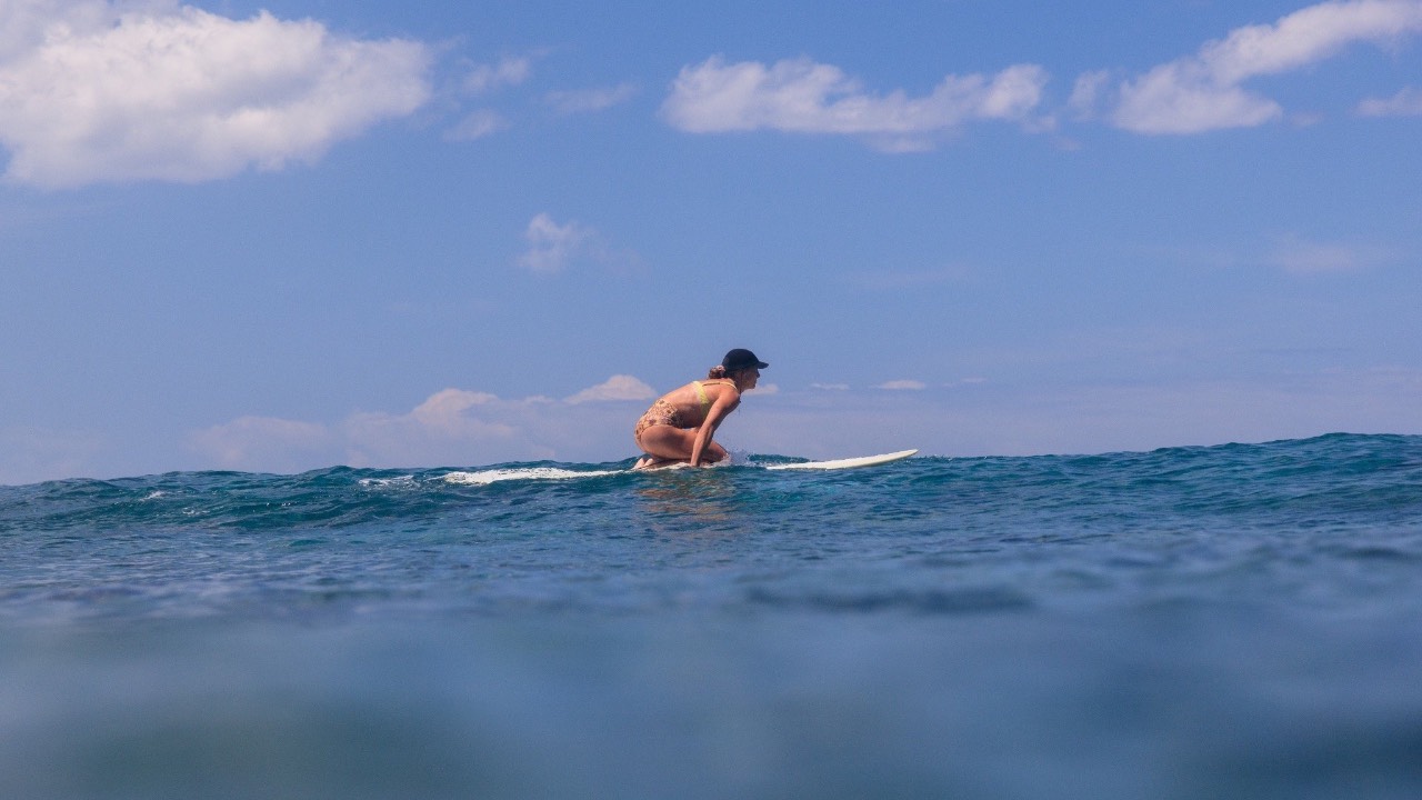 Woman riding a wave on a surfboard, crouched and focused in the ocean under a bright blue sky.