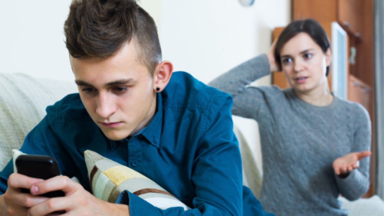 Teen boy focused on his phone with his back to his mom as she tries to engage, reflecting the quiet pressure and disconnection many families experience.