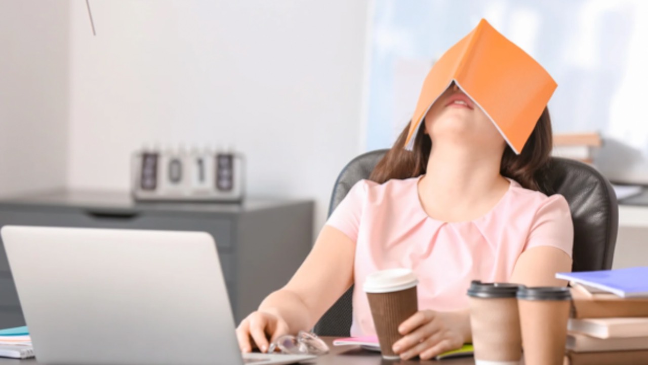 Overwhelmed mom working at a kitchen table with a laptop, three coffee cups, and a book covering her face