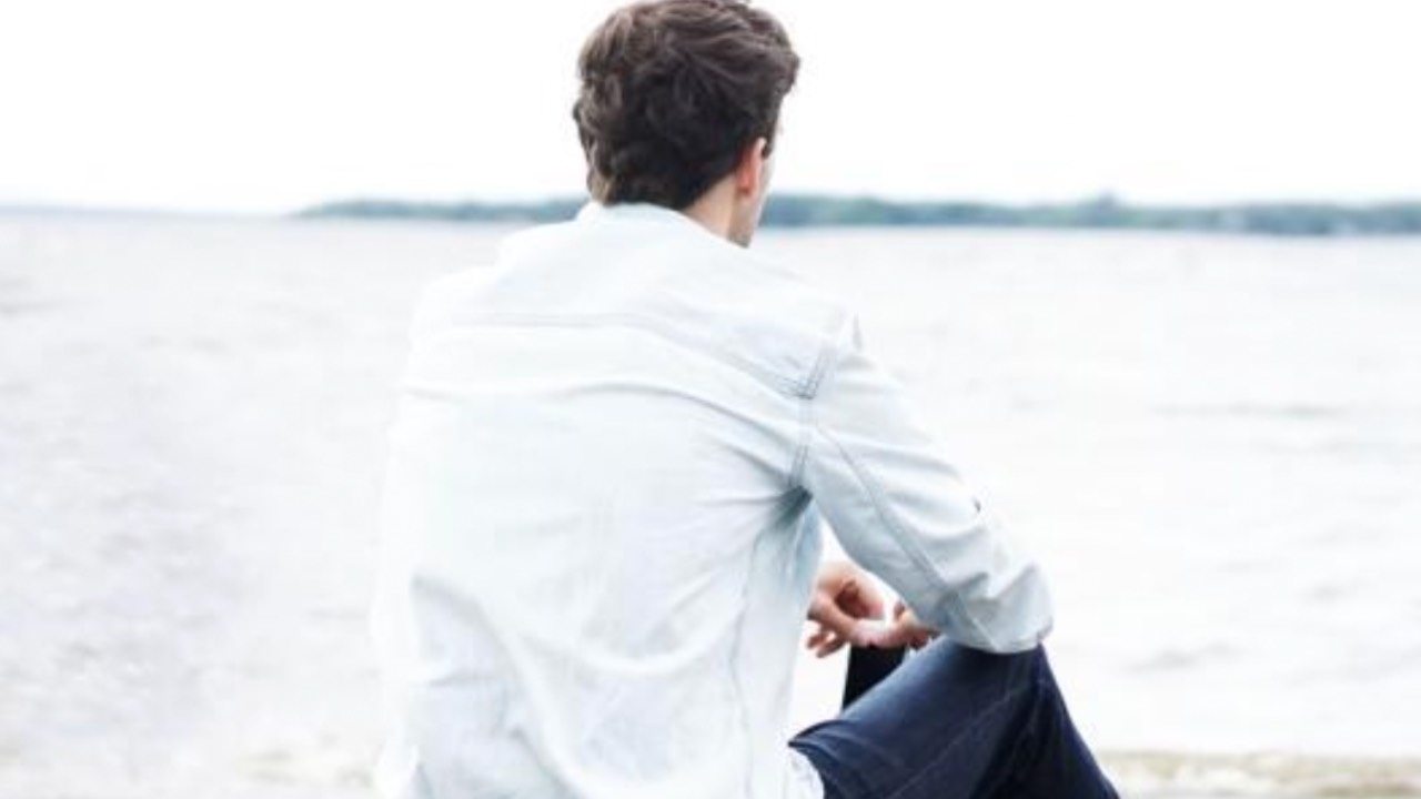 Young man sitting alone on the beach looking at the ocean, representing teen stress and pressure