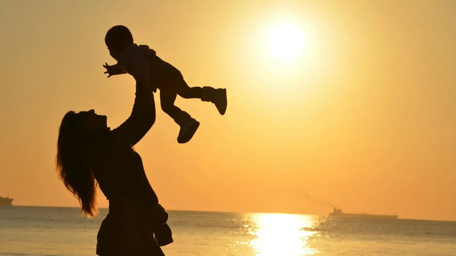 Silhouette of a mother lifting her baby in the air on the beach at sunset.