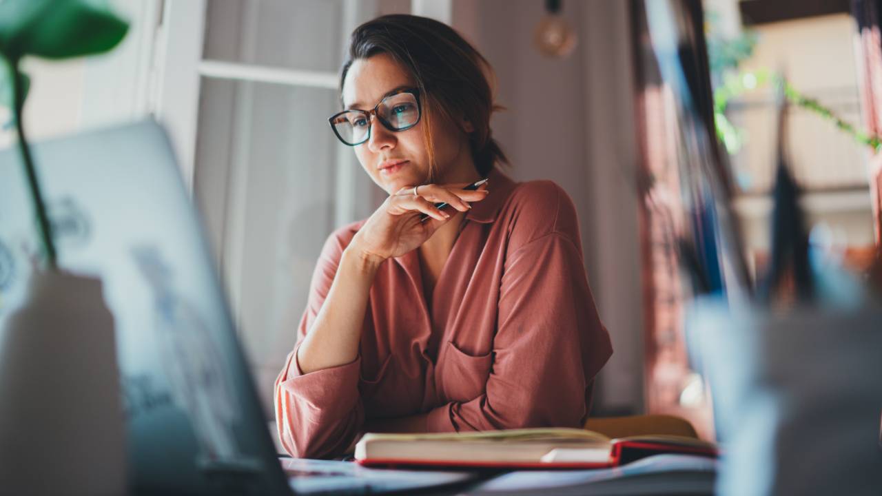 A woman wearing glasses sits at her desk with her laptop open, gently resting her hand on her chin as she looks thoughtfully at the screen. Soft morning light fills the room, creating a calm, reflective mood as she considers her next financial steps.