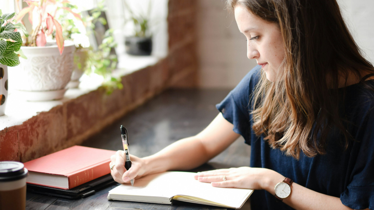 Woman journaling in a calm, sunlit café, symbolizing self-forgiveness, reflection, and taking a fresh start with her finances.