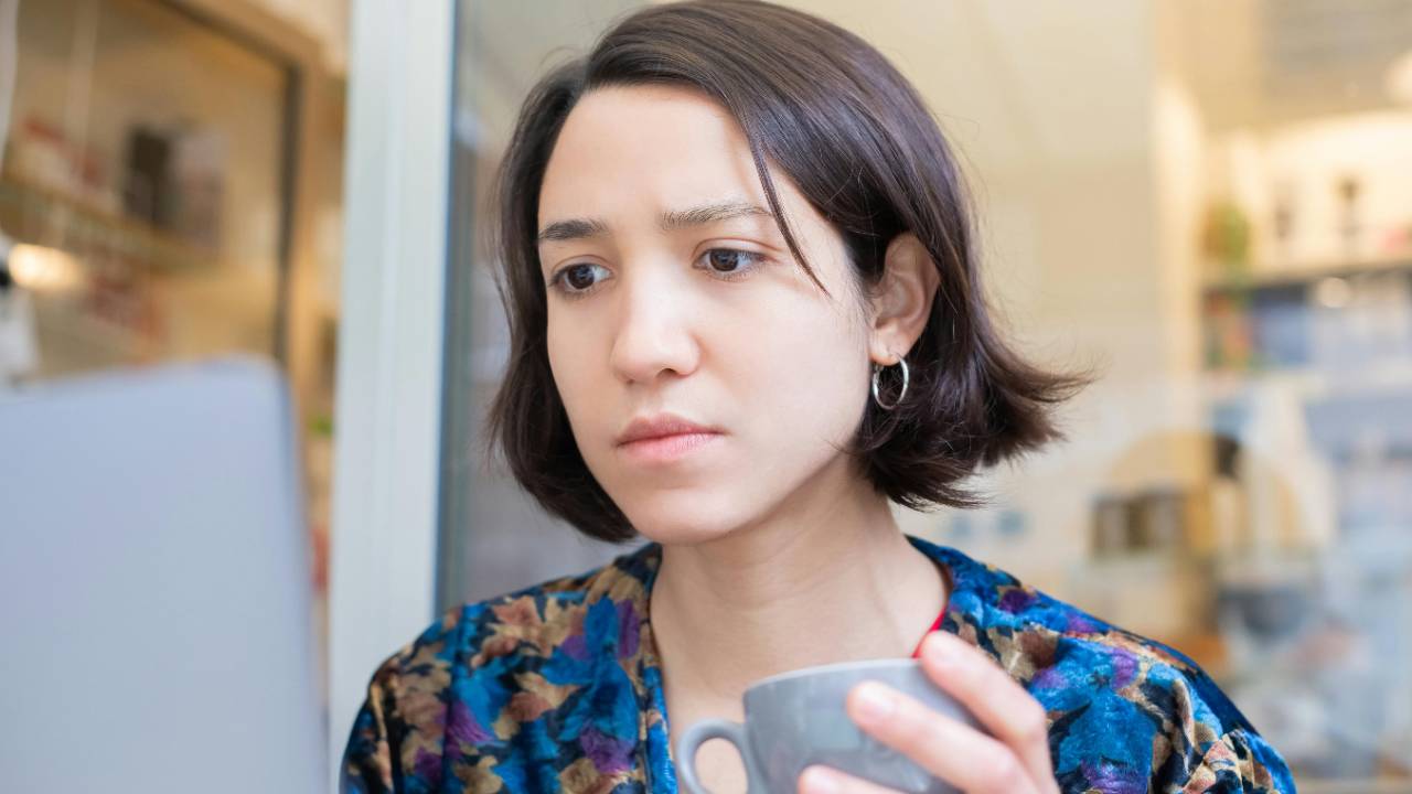 Woman sitting at a table holding a cup of coffee while looking at her laptop, pausing to face her finances with quiet focus and honesty.