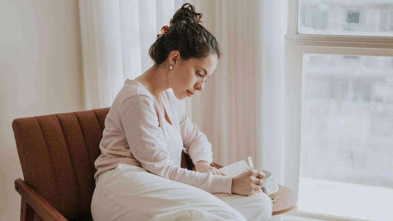 Woman journaling with a cup of coffee in soft morning light, creating a calm weekly money ritual to reduce financial stress.