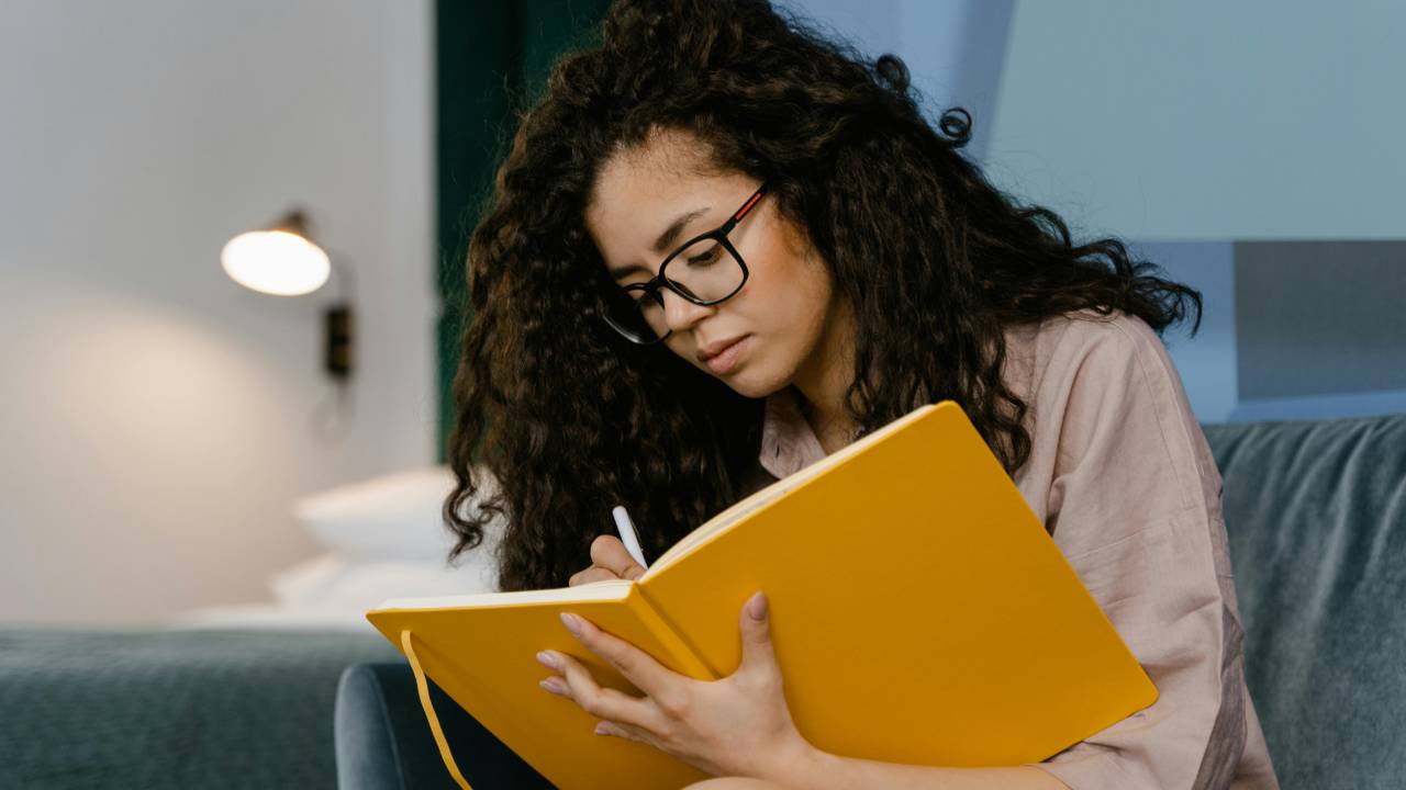 A woman sitting on a couch journaling in a yellow notebook, reflecting quietly during a calm weekly money check-in.