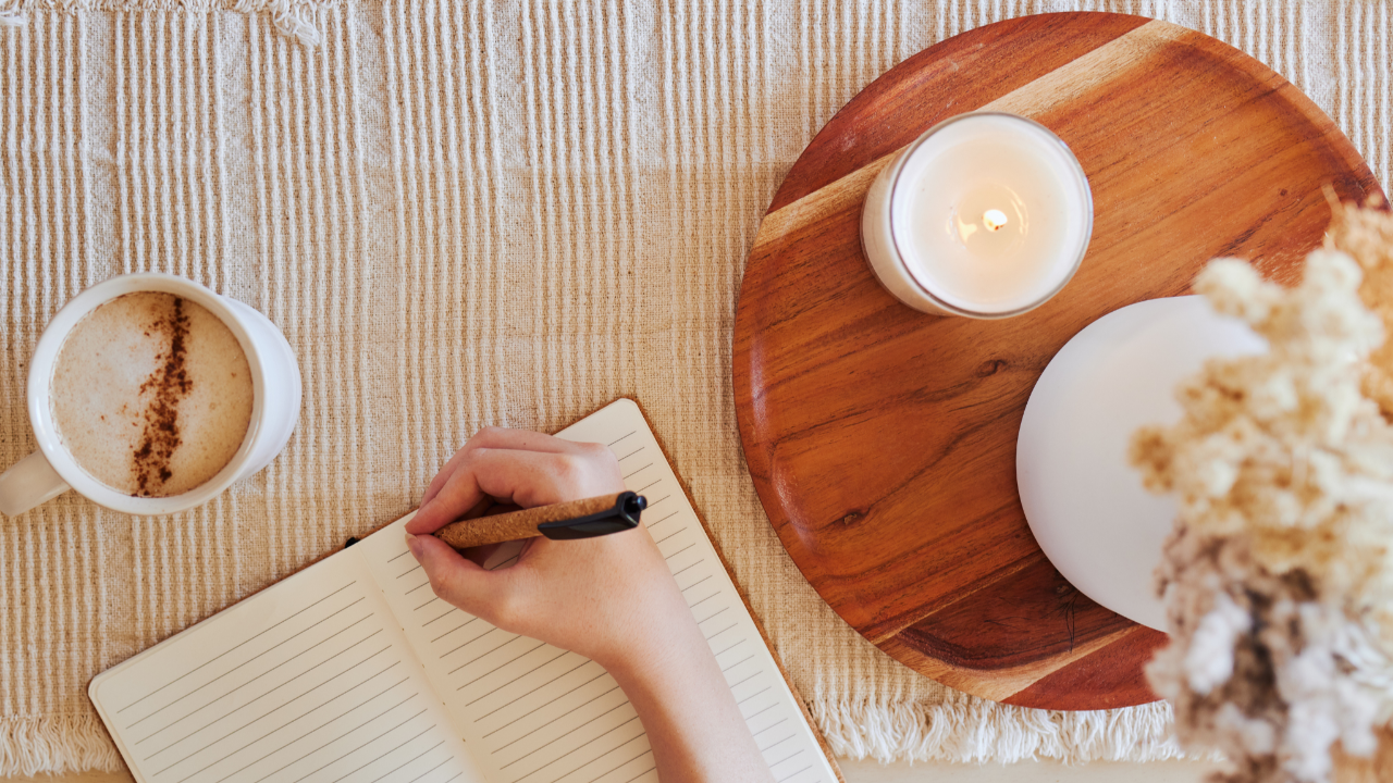Overhead view of a soft, calming workspace with a latte, a lit candle, and an open journal as a woman begins a gentle 10-minute weekly money check-in.