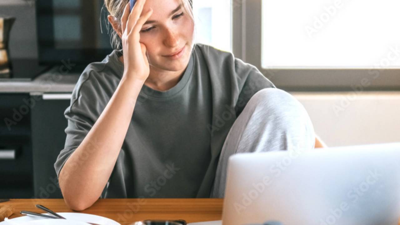 A woman sitting at her kitchen table, gently holding her head as she looks at her laptop, capturing the quiet moment she realizes it’s time to start over financially.