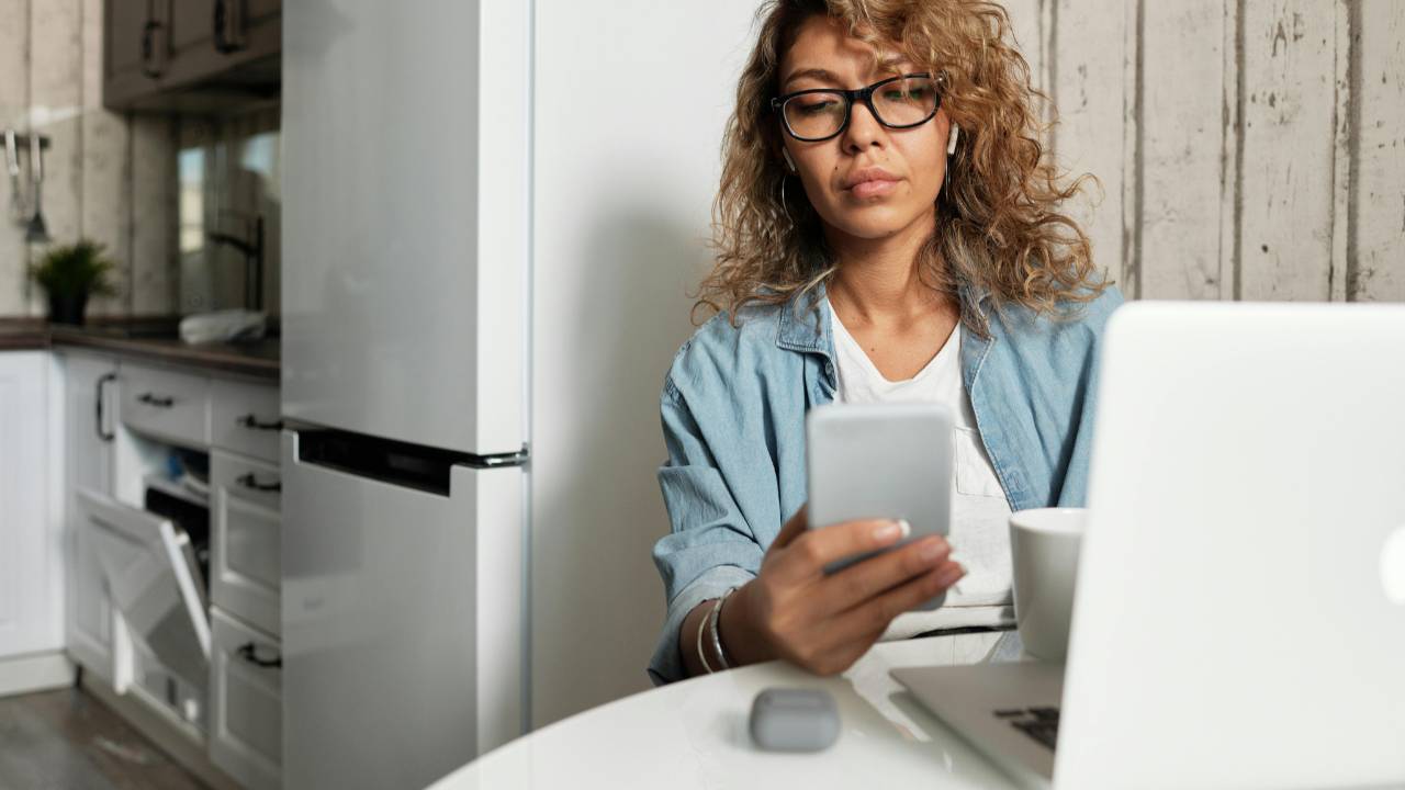 A woman sitting at her kitchen table looking worried as she checks her phone beside her laptop, reflecting the emotional weight of facing financial stress alone.