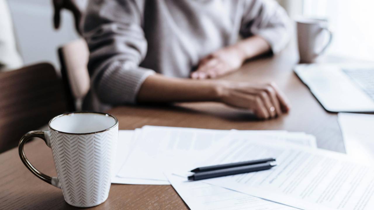 Woman sitting at a table with coffee and paperwork, calmly reviewing finances in a gentle, low-pressure way
