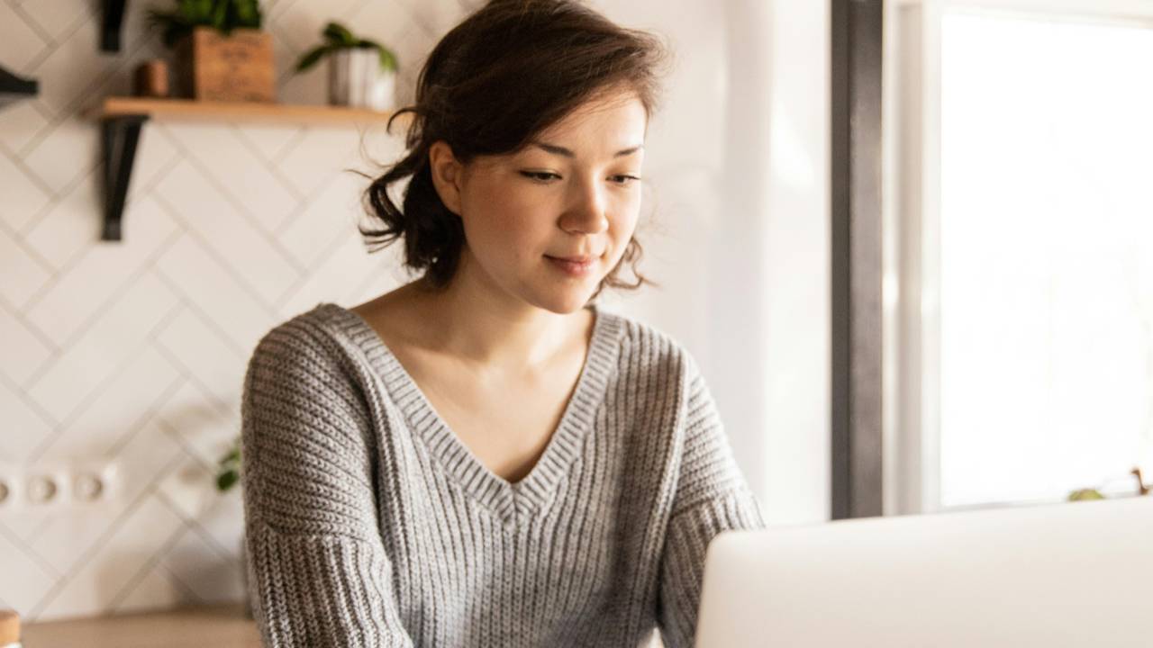 Woman sitting at her kitchen table using a laptop, calmly reviewing her finances in a warm, peaceful setting.
