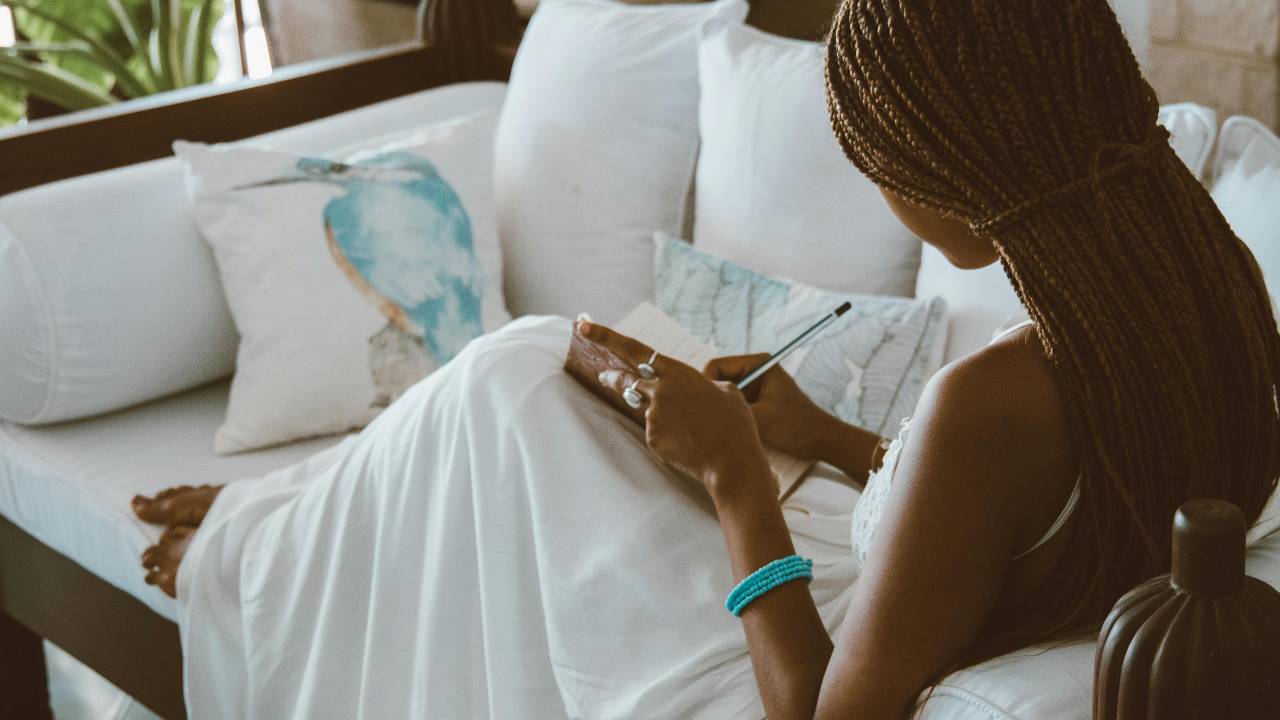 A woman journaling quietly on a couch in soft natural light, reflecting and releasing emotional weight around money.