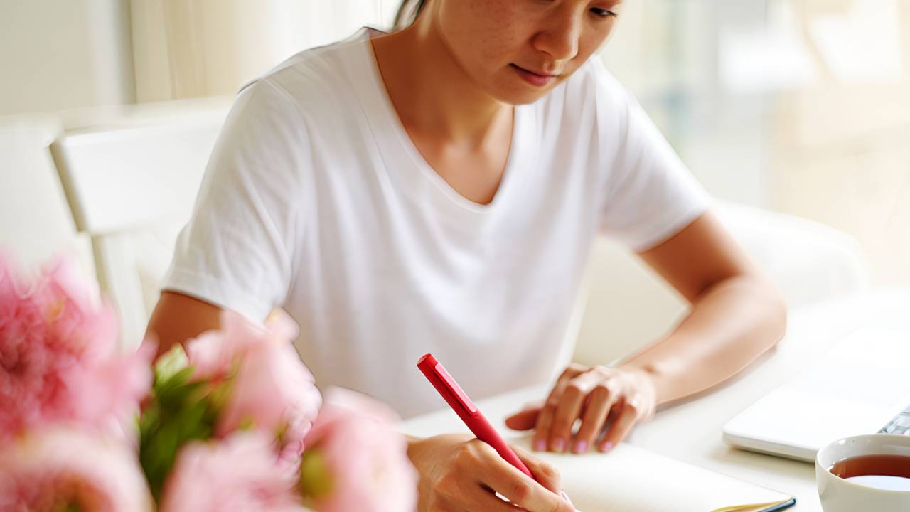 Woman sitting at a bright white desk, journaling with a red pen beside a cup of tea and pink flowers, creating a calm and reflective moment as she begins facing her finances.