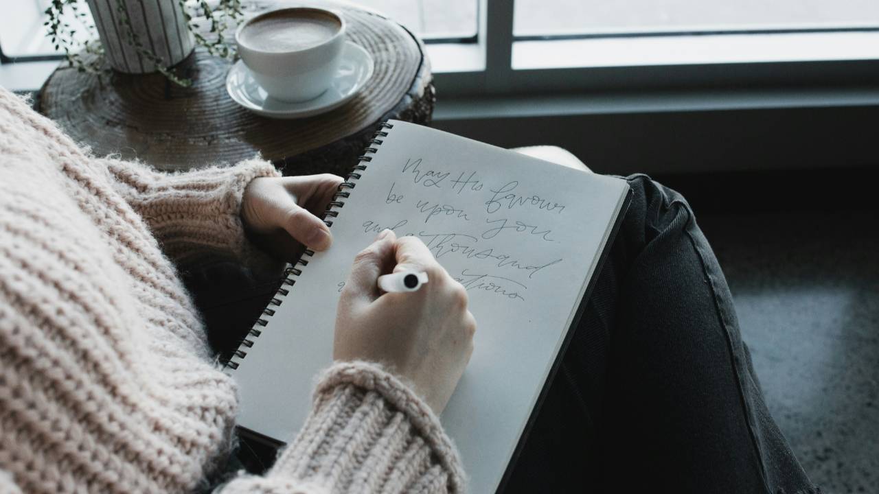 Woman journaling by a window with a cup of coffee, reflecting gently on finances and releasing shame while taking calm steps toward financial confidence.