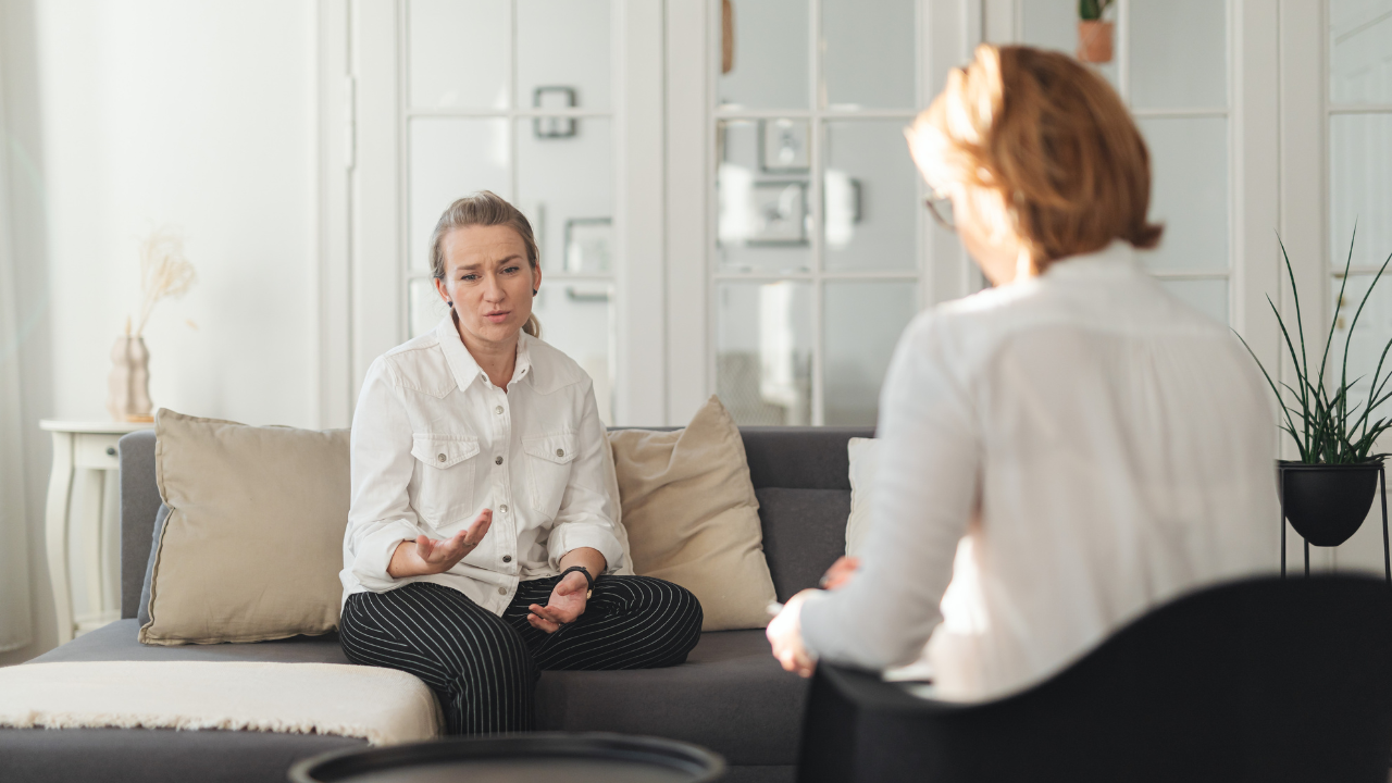 Una mujer sentada en un sofá gesticula mientras habla con una profesional que toma notas en un cuaderno durante una sesión de consultoría o terapia en una oficina luminosa