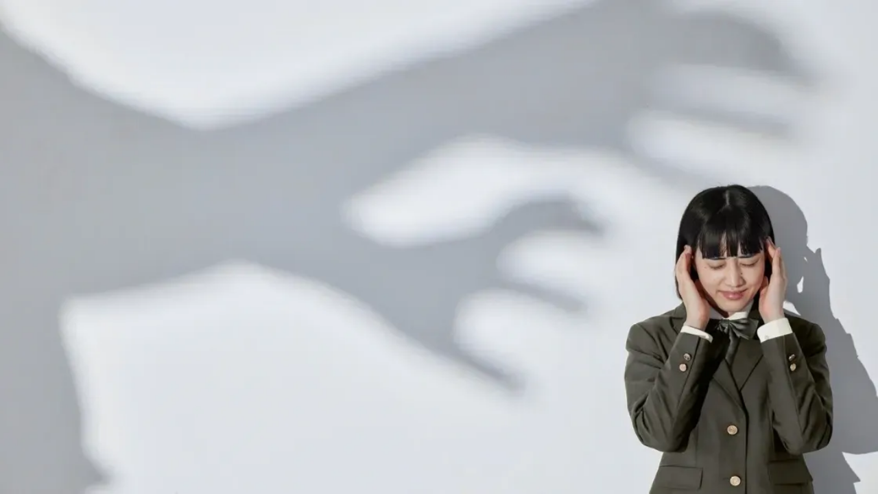 An overwhelmed woman stands against a wall, covering her ears with eyes closed.