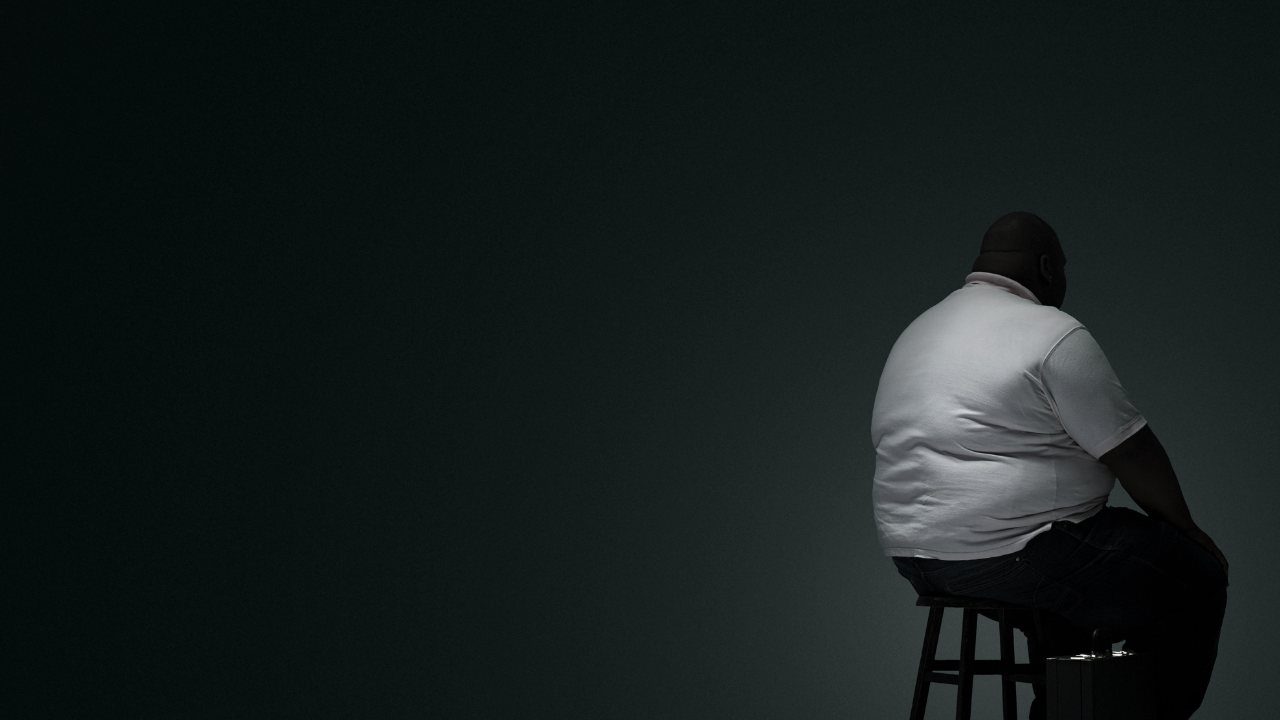 Large man sitting on a stool with his back turned in a dark room