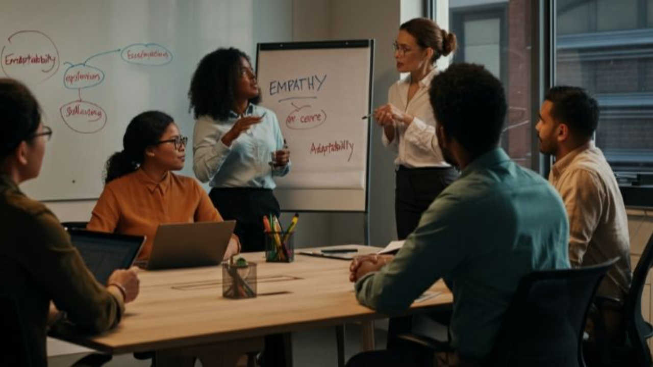 Two female presenters leading a professional workshop on empathy and adaptability, with a diverse group of attendees seated around a table in a modern office.