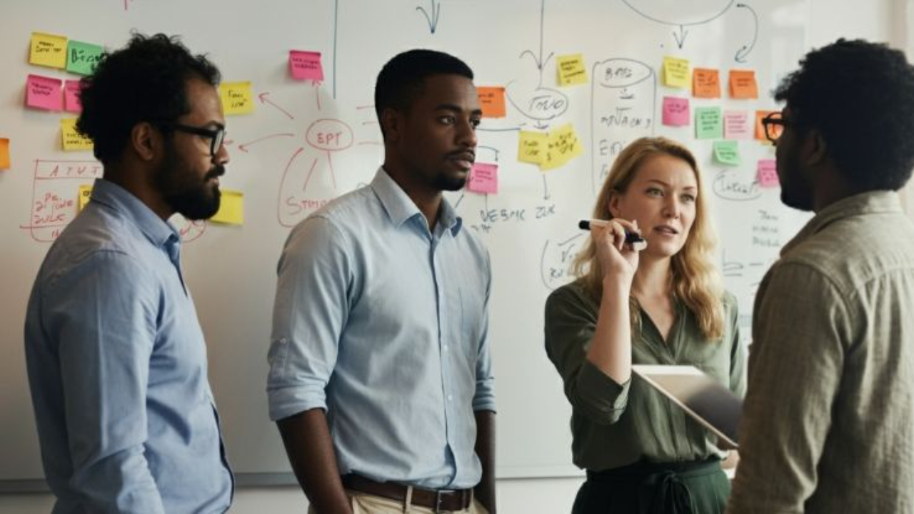 Four professionals collaborating in a brightly lit office, standing in front of a whiteboard filled with diagrams and colorful sticky notes, engaged in brainstorming.