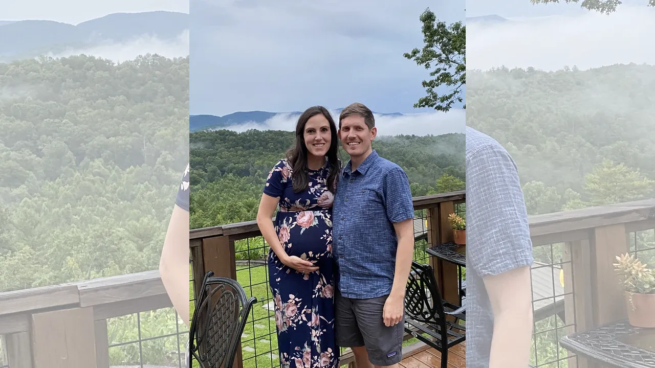 A couple stands together smiling for a photo. One partner is visibly pregnant, and both are posed warmly together in anticipation of their baby's arrival.