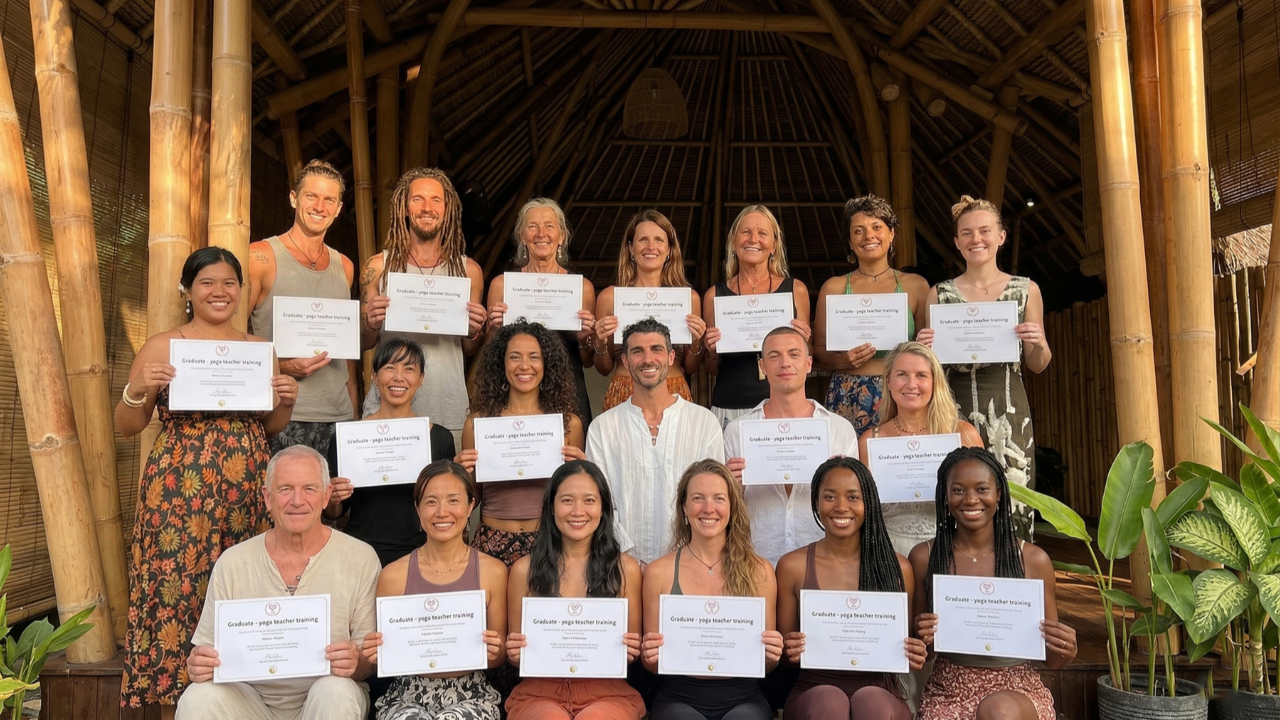 A diverse group of eighteen people holds identical "Graduate - yoga teacher training" certificates under a bamboo structure, smiling, celebrating, and surrounded by plants.