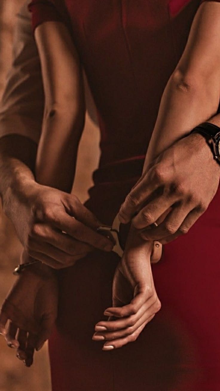 A man stands behind a woman in a red dress, holding her wrists together in front of her