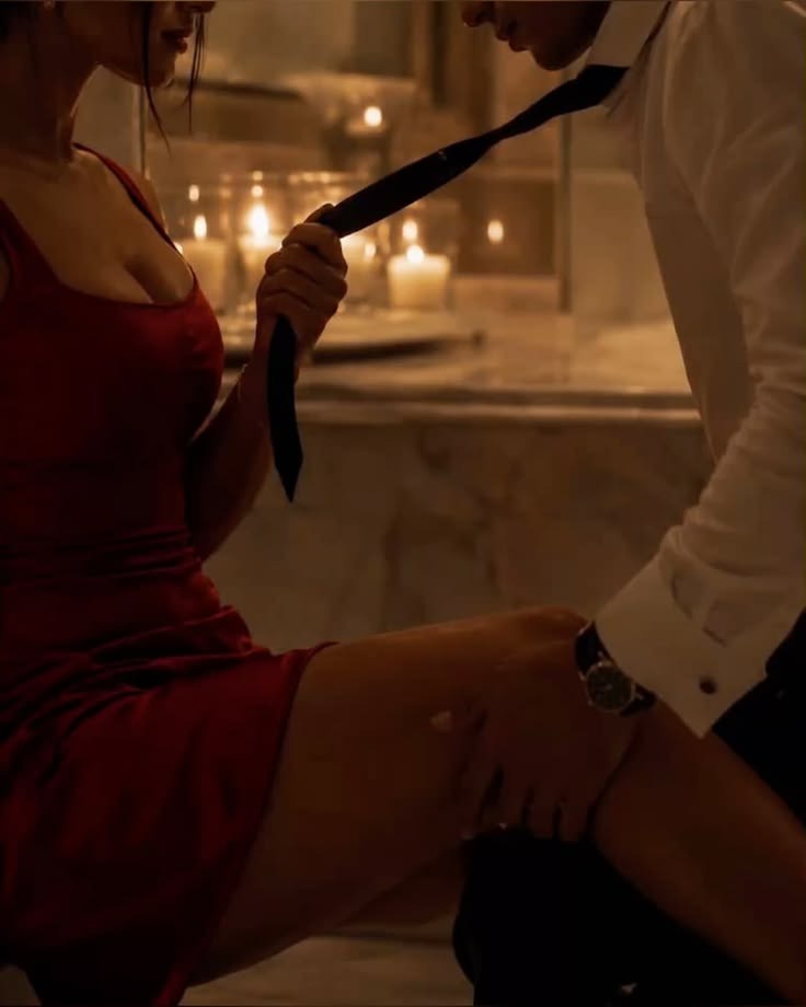 A woman in a red dress pulls her lover close by his tie in candlelight